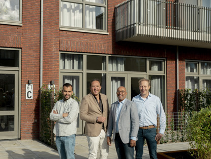 Vier mannen staan voor een modern appartementencomplex met grote ramen en een balkon, gekleed in casual en zakelijke kleding.