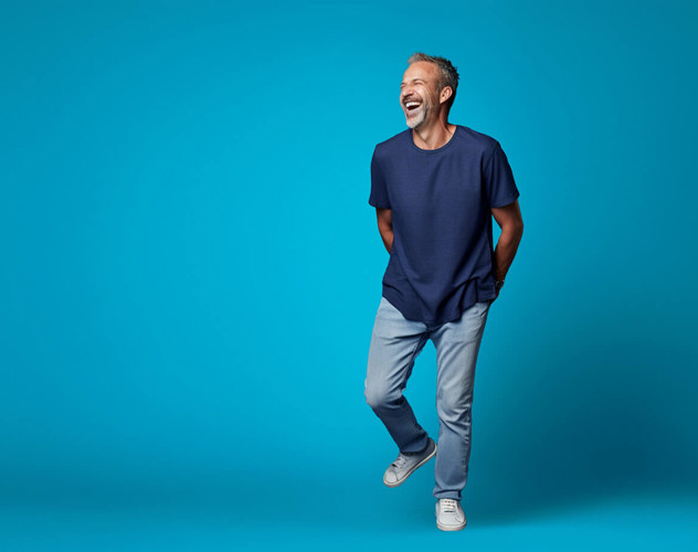 Man in een blauw T-shirt en lichte jeans, staand voor een felblauwe achtergrond.