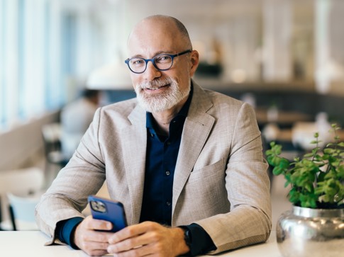 Een man in een lichte blazer zit aan een tafel met een smartphone in zijn hand, omgeven door een moderne interieur.