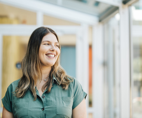 Vrouw met lang, golvend haar in een groene blouse, staande bij de glazen deuren van een modern gebouw.