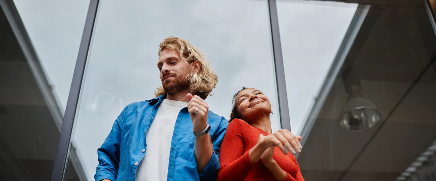 Twee mensen dansen blij voor een glazen gevel; de man draagt een blauw shirt en de vrouw een rode top.