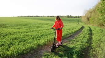 Vrouw in een felrode trainingsoutfit rijdt op een elektrische step over een smal pad in een groen, uitgestrekt veld.