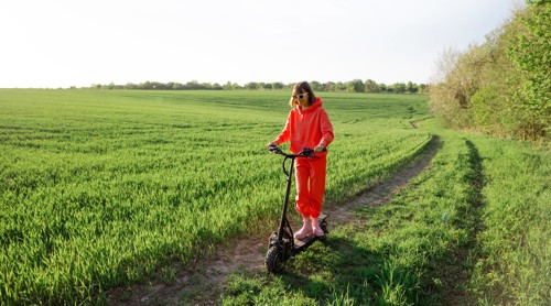 Vrouw in een felrode trainingsoutfit rijdt op een elektrische step over een smal pad in een groen, uitgestrekt veld.
