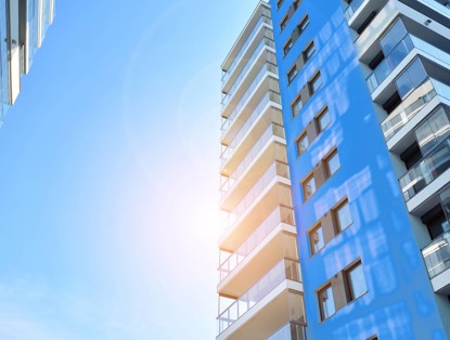 Hoog gebouw met glazen balkons en een blauwe gevel, tegen een heldere blauwe lucht met zon.