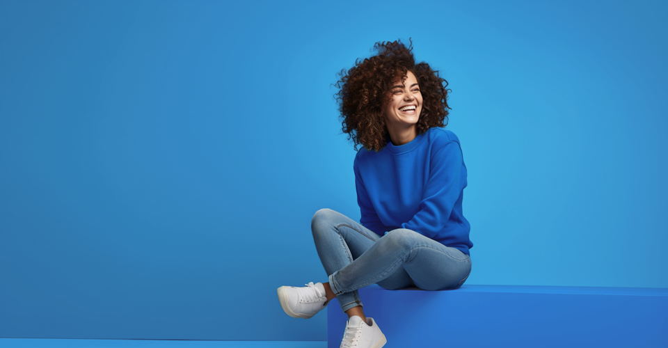 Een vrouw in een blauwe sweater zit op de grond met een blauwe achtergrond, glimlachend en met gekruiste benen.