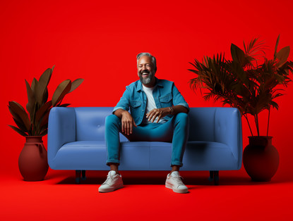 Man in een blauwe outfit zit ontspannen op een lichtblauwe bank, tegen een felrode achtergrond, omringd door groene planten.