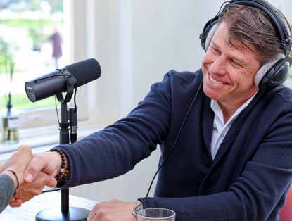 Twee mensen schudden elkaar de hand tijdens een podcastopname, terwijl een microfoon op de tafel zichtbaar is.