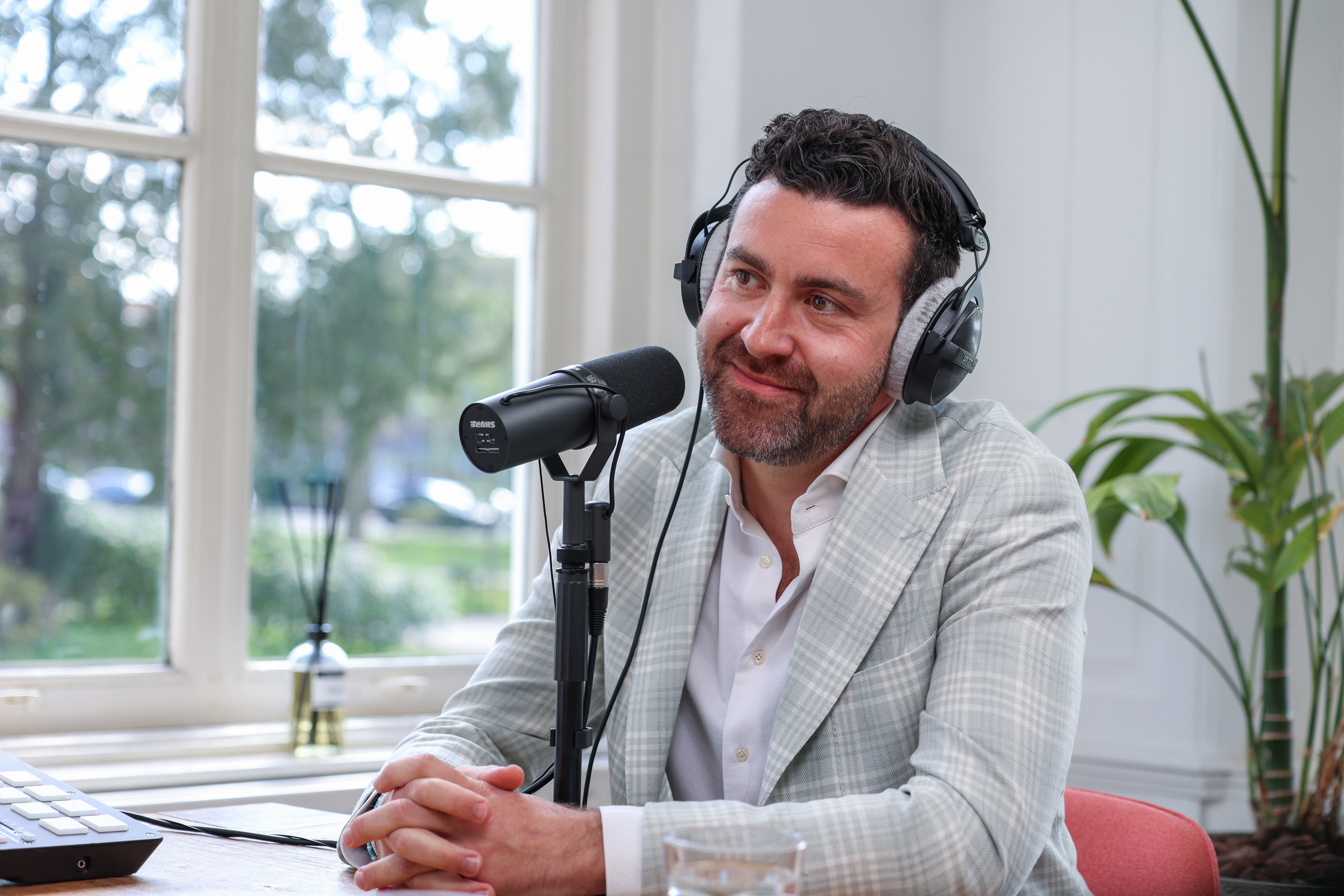 Man in een lichte blazer met een headset, zit aan een tafel voor een microfoon in een lichte, moderne ruimte.