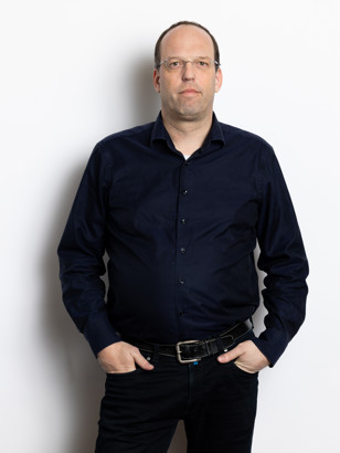 Paul Tromp in een donkerblauwe blouse met een zwarte riem, staand met één hand in zijn zak en de andere hand ontspannen naast zijn lichaam.