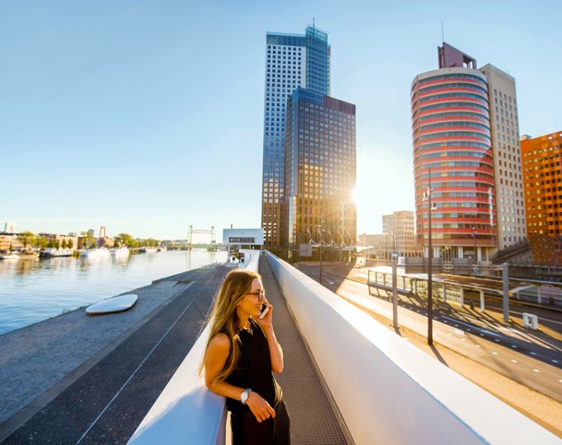 Een vrouw wandelt langs een moderne kade met gebouwen en een groot schip in de haven, terwijl het zonlicht de skyline verlicht.