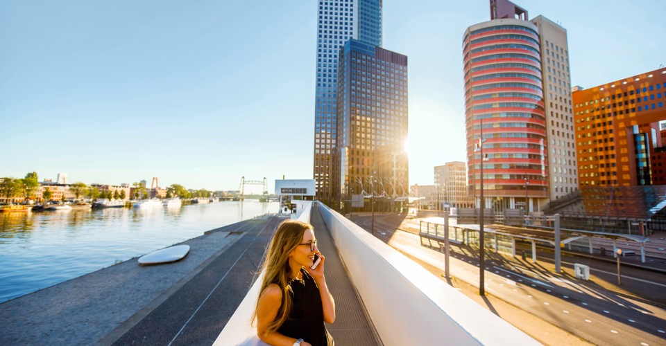 Een vrouw wandelt langs een moderne kade met gebouwen en een groot schip in de haven, terwijl het zonlicht de skyline verlicht.