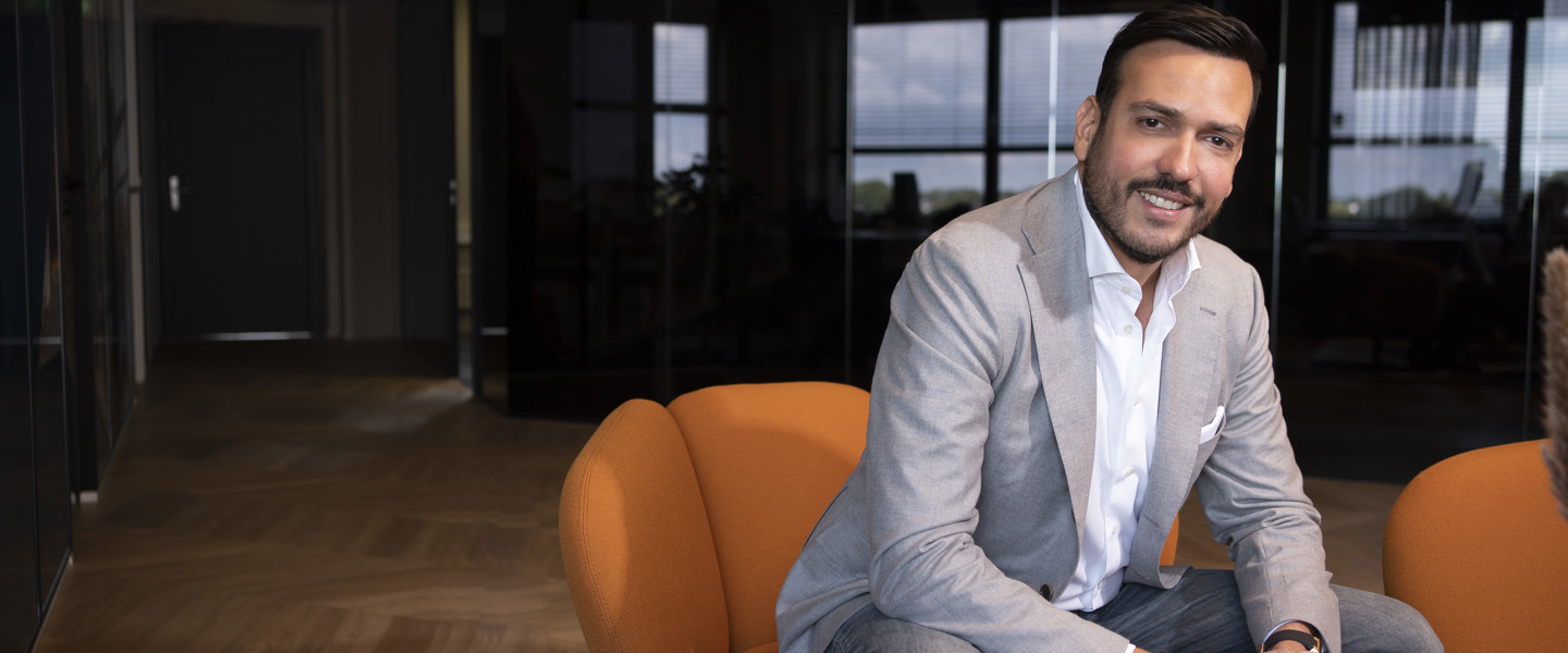 Een zakelijke Frederico Angius in een lichtgrijze blazer zit op een oranje fauteuil, met een moderne kantooromgeving op de achtergrond.