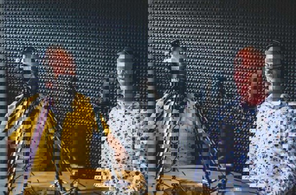 Twee mannen zitten aan een tafel in een opnamestudio, met een microfoon voor zich en een koptelefoon op.