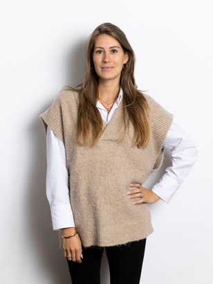 Ella Van Imhoff in een beige oversized trui met een witte blouse eronder, poseert met haar hand op haar heup tegen een witte achtergrond.