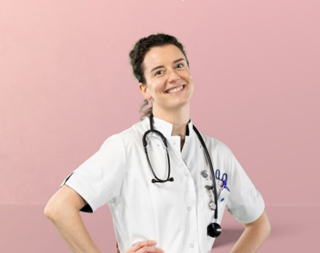 Zorgprofessional in witte labjas met stethoscoop om de nek, staand met handen op de heupen tegen een roze achtergrond.