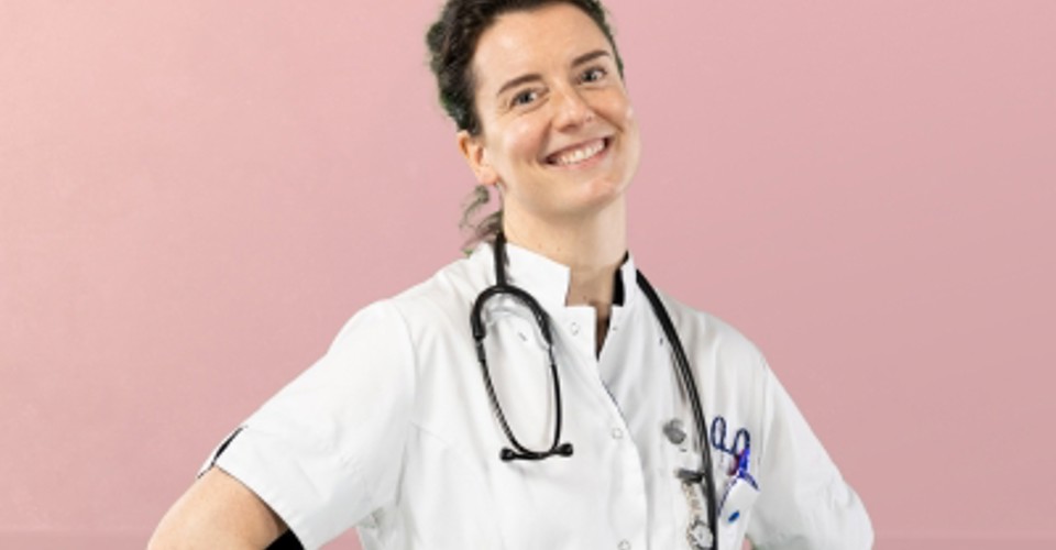 Zorgprofessional in witte labjas met stethoscoop om de nek, staand met handen op de heupen tegen een roze achtergrond.