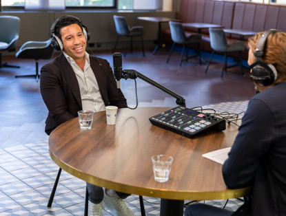 Twee mannen zitten aan een ronde tafel in een moderne ruimte; de ene spreekt in een microfoon terwijl de andere aandachtig luistert.