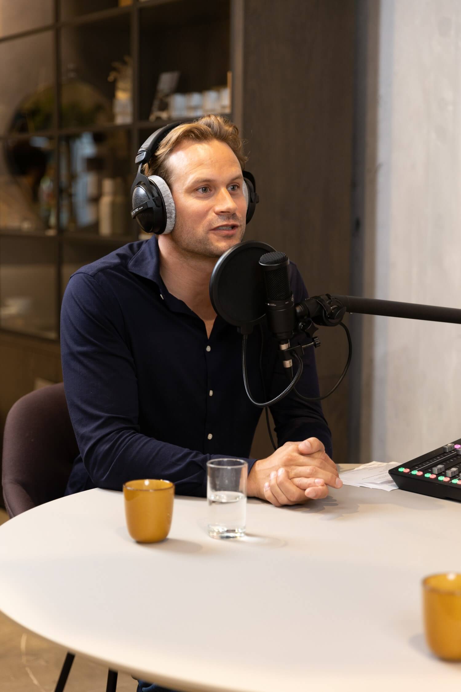 Een man met een hoofdtelefoon zit achter een microfoon in een sfeervolle studio.