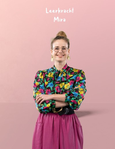 Leerkracht Mira in een kleurrijke blouse en paarse rok, staand voor een roze achtergrond met een zelfverzekerde uitstraling.