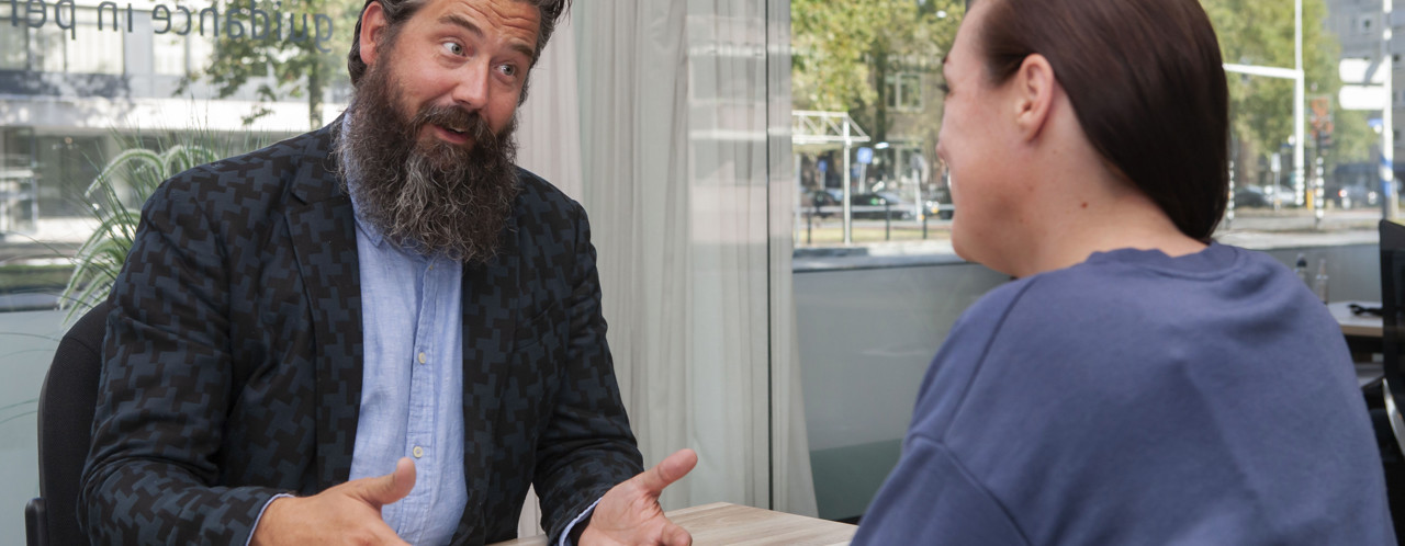 Een man in een blazer bespreekt iets met een vrouw in een casual outfit, in een kantoor met grote ramen.