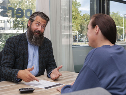 Een man in een blazer bespreekt iets met een vrouw in een casual outfit, in een kantoor met grote ramen.