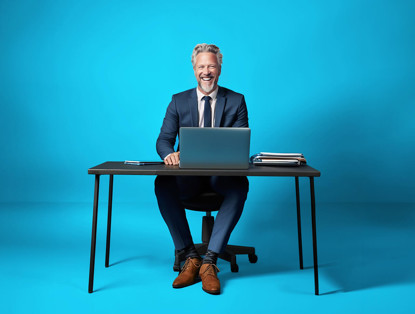Zittende man in een pak achter een bureau met een laptop, tegen een heldere blauwe achtergrond.