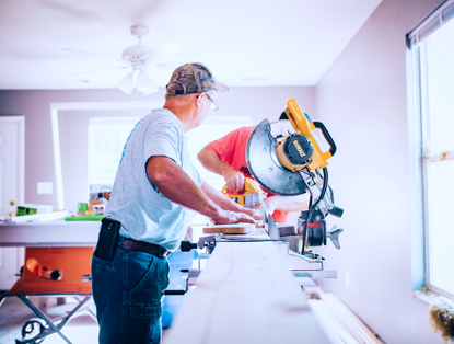 Twee mannen zijn aan het werk in een lichte kamer; de ene zaagt een plank met een zaagmachine terwijl de ander toekijkt.