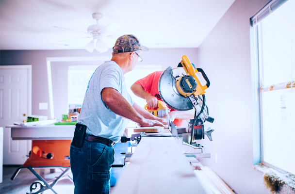 Twee mannen zijn aan het werk in een lichte kamer; de ene zaagt een plank met een zaagmachine terwijl de ander toekijkt.