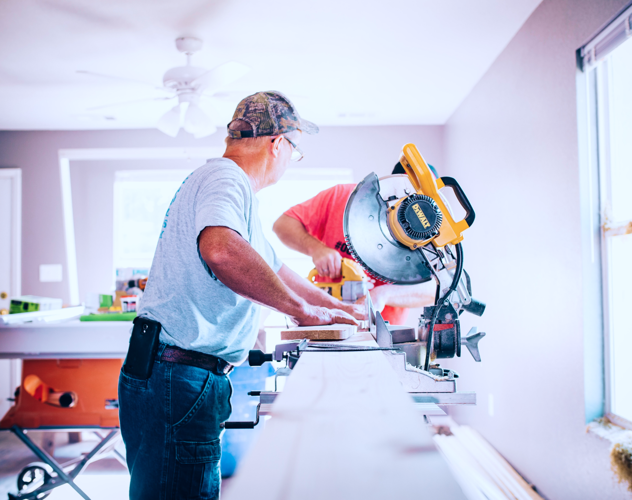 Twee mannen zijn aan het werk in een lichte kamer; de ene zaagt een plank met een zaagmachine terwijl de ander toekijkt.