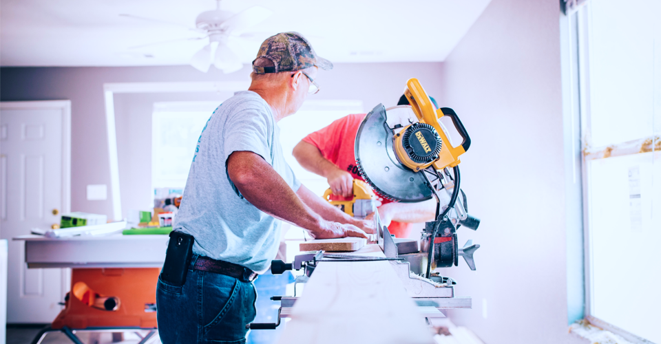 Twee mannen zijn aan het werk in een lichte kamer; de ene zaagt een plank met een zaagmachine terwijl de ander toekijkt.