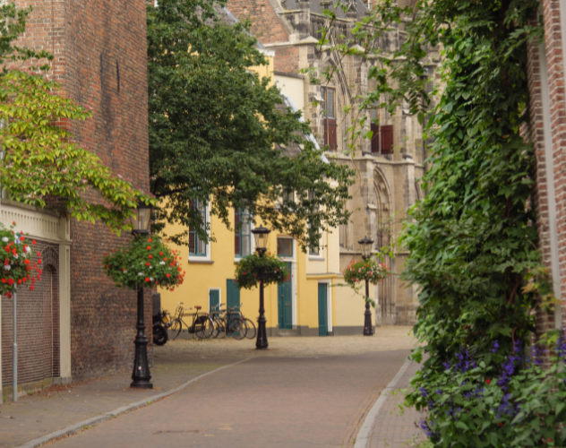 Rustige straat met gele en groene gebouwen, lantaarns en bloemen op de muren; geparkeerde fietsen.