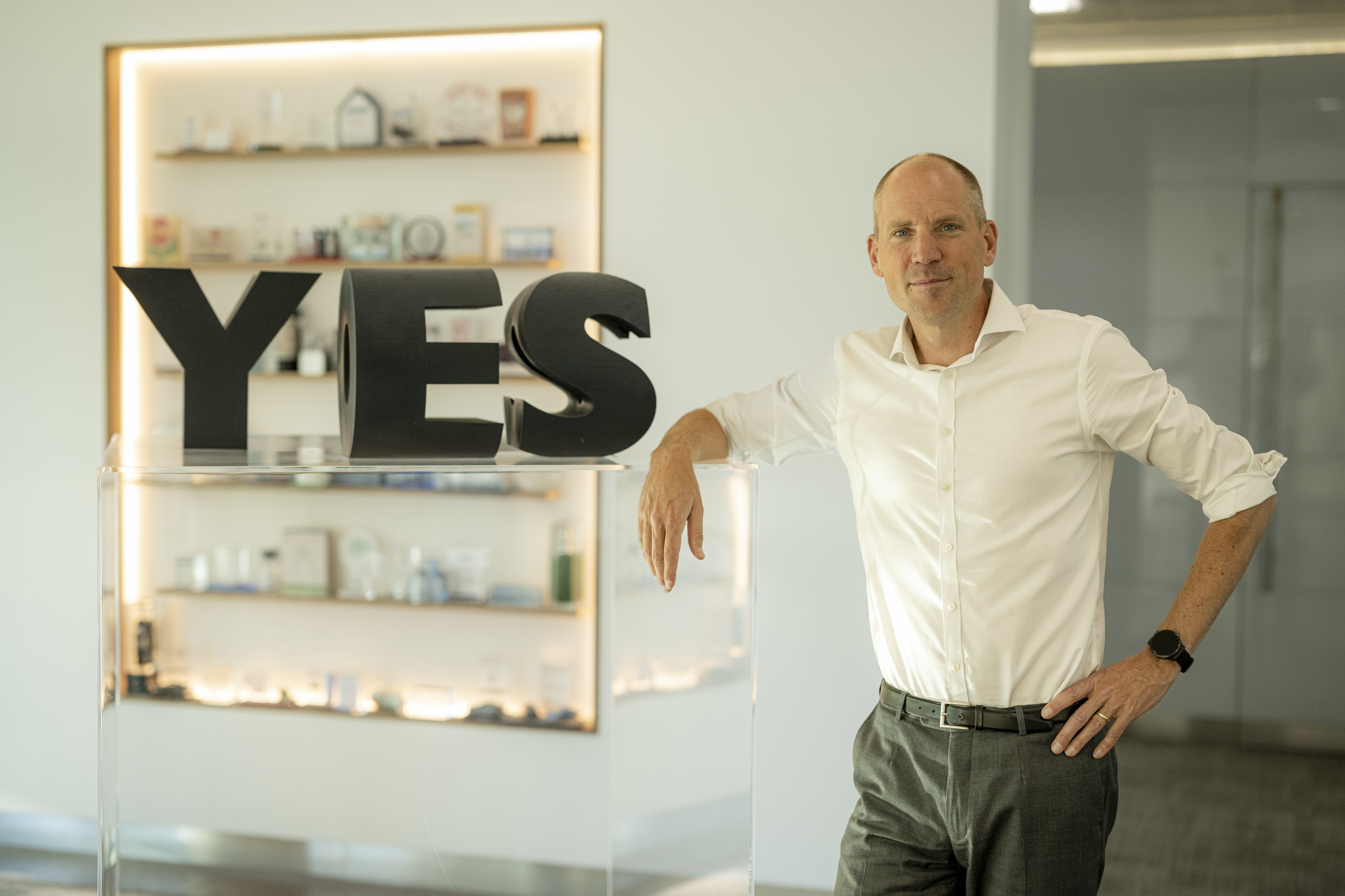 Een man in een witte blouse leunt op een transparante balie met grote letters 'ES' ernaast. Op de achtergrond zijn verschillende objecten op een plank zichtbaar.