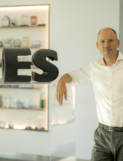 Een man in een witte blouse leunt op een transparante balie met grote letters 'ES' ernaast. Op de achtergrond zijn verschillende objecten op een plank zichtbaar.