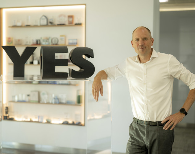 Een man in een witte blouse leunt op een transparante balie met grote letters 'ES' ernaast. Op de achtergrond zijn verschillende objecten op een plank zichtbaar.