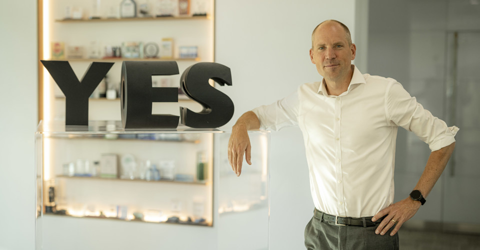 Een man in een witte blouse leunt op een transparante balie met grote letters 'ES' ernaast. Op de achtergrond zijn verschillende objecten op een plank zichtbaar.