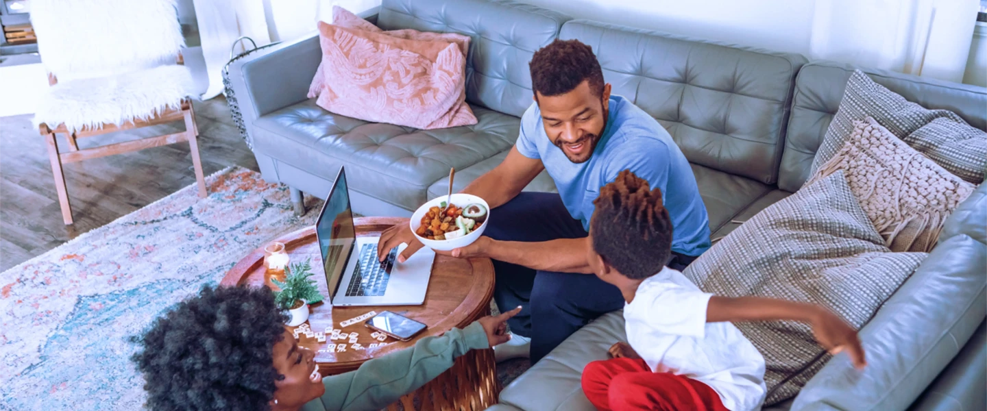 Man zit op een bank met een laptop, terwijl twee kinderen naast hem spelen en hij een schaal met eten vasthoudt.