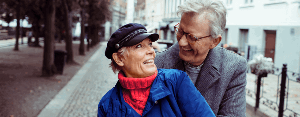 Een oudere vrouw in een blauwe jas en een oudere man in een lichte jas zitten op een straat omringd door bomen en gebouwen op de achtergrond.