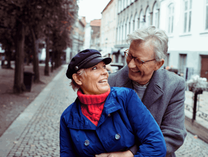 Een oudere vrouw in een blauwe jas en een oudere man in een lichte jas zitten op een straat omringd door bomen en gebouwen op de achtergrond.