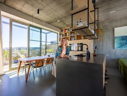 Een glimlachende vrouw leunt op een moderne keukenbar in een lichte, open woonkamer met een eettafel en grote ramen.