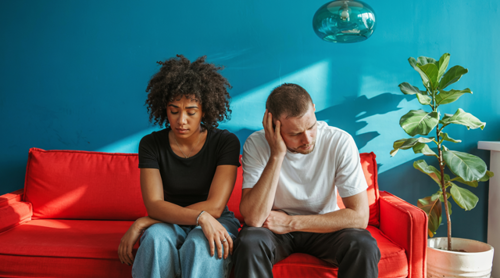 Een vrouw in een zwarte top en een man in een wit T-shirt zitten op een rode bank, met een groene plant op de achtergrond.