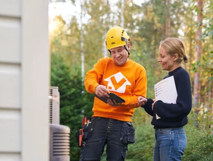 Een man in een oranje sweatshirt met een logo toont iets op een smartphone aan een vrouw met een bloknote, in een bosrijke omgeving.