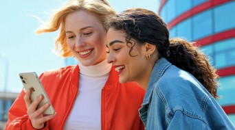 Twee jonge vrouwen bekijken samen een smartphone buiten, met een modern gebouw op de achtergrond.