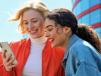 Twee jonge vrouwen bekijken samen een smartphone buiten, met een modern gebouw op de achtergrond.