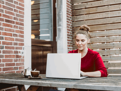 Een persoon in een rode trui werkt aan een laptop aan een houten tafel buiten, met een koffiezetapparaat en een kopje naast zich.