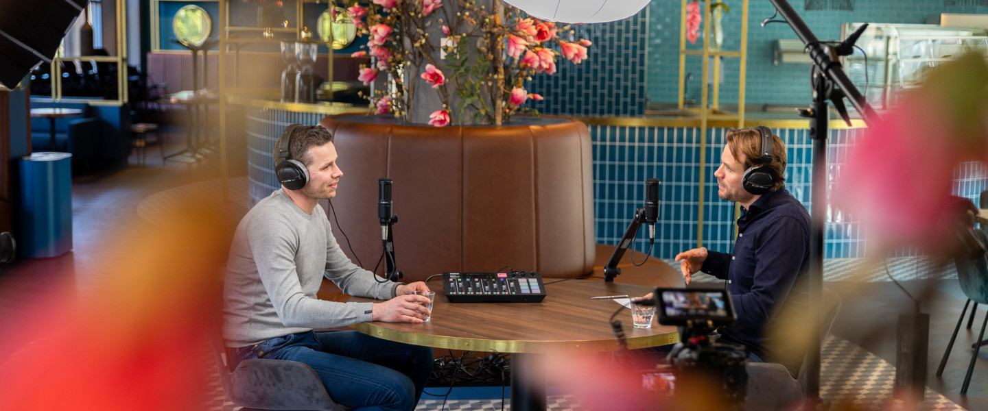 Twee mannen zitten tegenover elkaar in een café, omringd door microfoons en opnameapparatuur op de tafel.