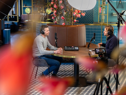 Twee mannen zitten tegenover elkaar in een café, omringd door microfoons en opnameapparatuur op de tafel.