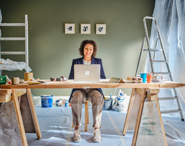 Een persoon werkt achter een laptop aan een tafel vol schildermaterialen in een gerenoveerde ruimte. Aan de muur hangen drie schilderijtjes.