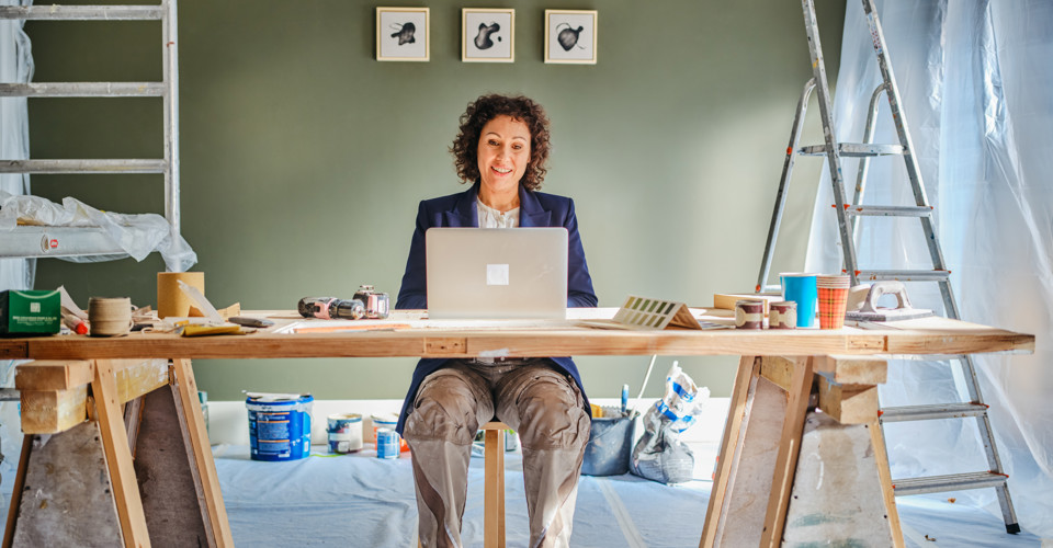 Een persoon werkt achter een laptop aan een tafel vol schildermaterialen in een gerenoveerde ruimte. Aan de muur hangen drie schilderijtjes.