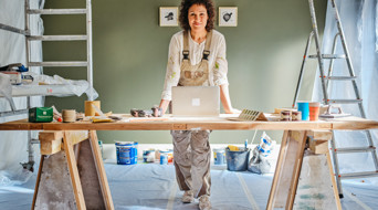 Een man in werkkleding staat aan een houten werktafel met een laptop, omringd door schildersmaterialen en een ladder.