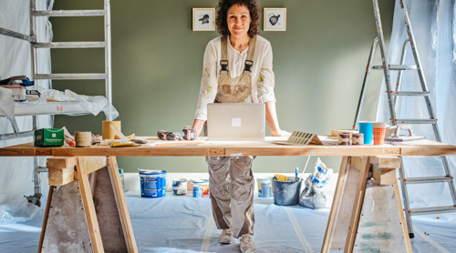 Een man in werkkleding staat aan een houten werktafel met een laptop, omringd door schildersmaterialen en een ladder.